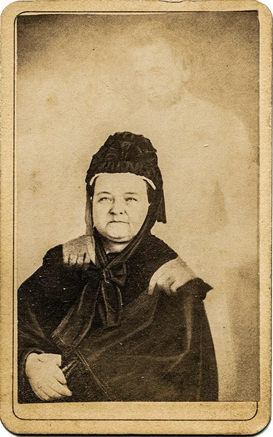 A faded carte-de-visite photograph of Mary Todd Lincoln in black mourning dress and bonnet, seated, with the pale, transparent figure of Abraham Lincoln standing behind her, his hands resting on her shoulders.